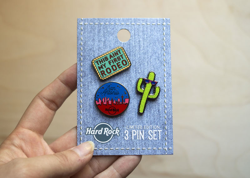 A hand holding a backing card with 3 enamel pins attached. 